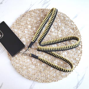 May include: A black smartphone lies on a woven placemat. A black and tan lanyard with a silver clasp is attached to the phone. The lanyard is made of woven cord. The placemat is circular.