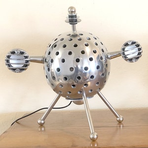 May include: A unique, silver-colored table lamp with a spherical body perforated with numerous small holes. The lamp has three legs and two side extensions, each with a spherical, caged light. A small antenna-like element sits atop the sphere.