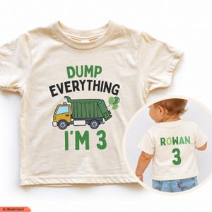 May include: A cream-colored t-shirt featuring a green and yellow dump truck graphic with the text DUMP EVERYTHING I'M 3. A circular inset shows the back of the shirt with the name ROWAN and the number 3 printed in green.