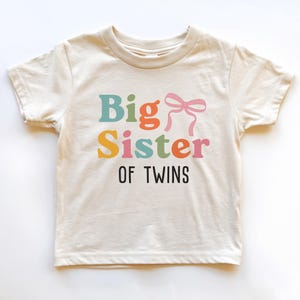 May include: A cream-coloured short-sleeve t-shirt featuring the text Big Sister in multi-coloured letters, a pink bow graphic, and the text OF TWINS in black lettering below. The shirt has a crew neckline and a standard straight hemline design.