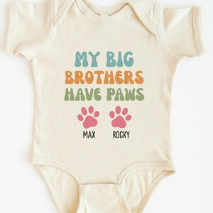 May include: Cream-colored baby bodysuit with the text "MY BIG BROTHERS HAVE PAWS" in teal, orange, and green. Below the text are two pink paw prints with the names "MAX" and "ROCKY" printed underneath.