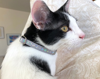 May include: A black and white cat with yellow eyes wearing a gray sparkly collar. The cat is looking to the right and is sitting on a patterned fabric.