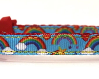 Op de afbeelding: Een blauwe kattenhalsband met een regenboog- en wolkenpatroon. De halsband heeft een rode bloemvormige gesp, een zilveren bel en een metalen lus. De halsband is ongeveer 30 cm lang.
