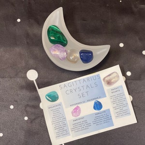 May include: A set of crystals for the Sagittarius zodiac sign. The crystals are arranged in a white crescent moon-shaped dish. The crystals are amethyst, smoky quartz, sodalite, and malachite. The crystals are labeled with their names and their astrological meanings.