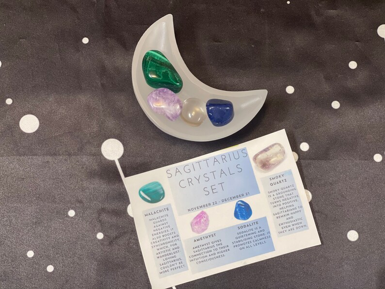 May include: A set of crystals for the Sagittarius zodiac sign. The crystals are arranged in a white crescent moon-shaped dish. The crystals are amethyst, smoky quartz, sodalite, and malachite. The crystals are labeled with their names and their astrological meanings.