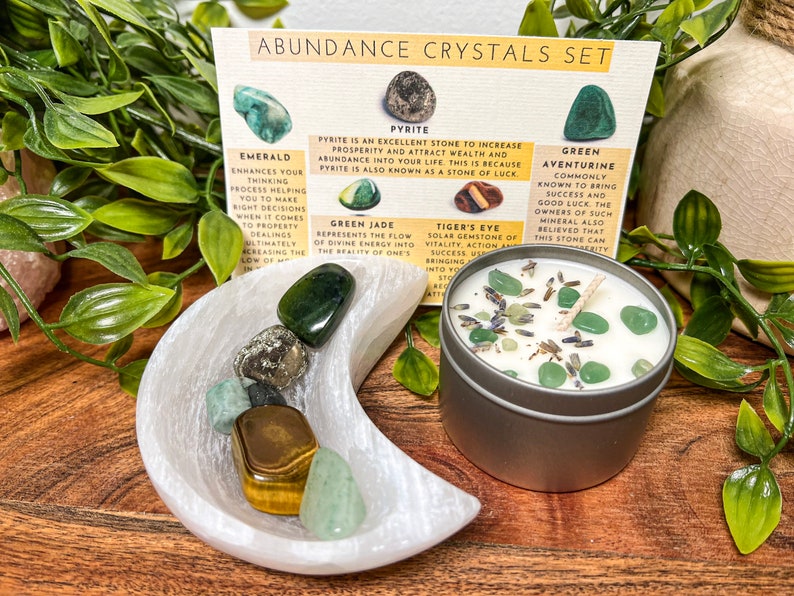 May include: An abundance crystals set featuring a variety of gemstones, including emerald, pyrite, and green aventurine, displayed with a candle and a card with descriptions. The set is arranged on a wooden surface.