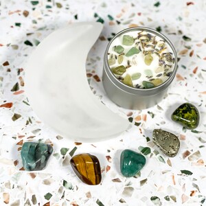 K&ouml;nnte beinhalten: Ein mattwei&szlig;er Halbmond, eine kleine silberne Kerzendose und mehrere Edelsteine sind auf einer gesprenkelten Oberfl&auml;che angeordnet. Die Kerze ist mit gr&uuml;nen Steinen und Lavendel bedeckt. Zu den Edelsteinen geh&ouml;ren Smaragd, Tigerauge und Pyrit.