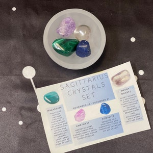 May include: A set of crystals for Sagittarius, November 22 - December 21. The set includes smoky quartz, amethyst, sodalite, malachite, and clear quartz. The crystals are arranged in a white bowl on a black background.