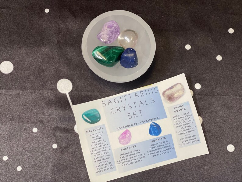 May include: A set of crystals for Sagittarius, November 22 - December 21. The set includes smoky quartz, amethyst, sodalite, malachite, and clear quartz. The crystals are arranged in a white bowl on a black background.
