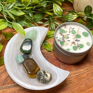 May include: A silver tin candle with a white wax base, green stones, and a wick. A white crescent moon-shaped dish holds various polished gemstones. Green foliage surrounds the objects, all set on a wooden surface.