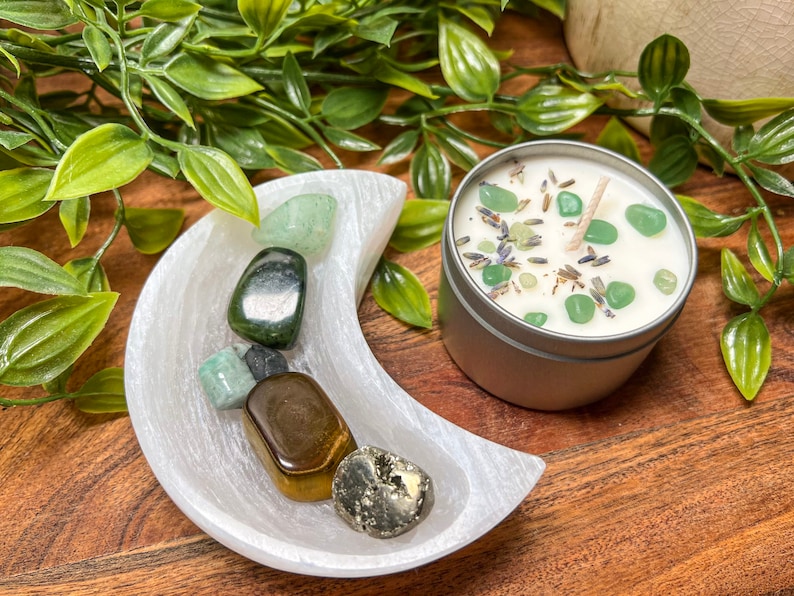 May include: A silver tin candle with a white wax base, green stones, and a wick. A white crescent moon-shaped dish holds various polished gemstones. Green foliage surrounds the objects, all set on a wooden surface.