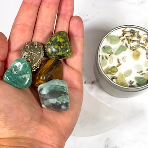 K&ouml;nnte beinhalten: Eine Sammlung von getrommelten Edelsteinen in verschiedenen Farben, darunter Gr&uuml;n, Braun und Gold, die in einer Hand gehalten werden. Eine kleine, silberne Kerzendose ist mit wei&szlig;em Wachs, gr&uuml;nen Steinen und getrocknetem Lavendel gef&uuml;llt. Die Artikel sind auf einer wei&szlig;en Oberfl&auml;che angeordnet.