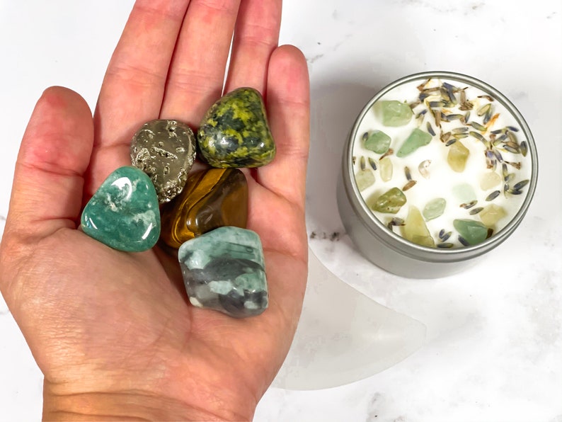 K&ouml;nnte beinhalten: Eine Sammlung von getrommelten Edelsteinen in verschiedenen Farben, darunter Gr&uuml;n, Braun und Gold, die in einer Hand gehalten werden. Eine kleine, silberne Kerzendose ist mit wei&szlig;em Wachs, gr&uuml;nen Steinen und getrocknetem Lavendel gef&uuml;llt. Die Artikel sind auf einer wei&szlig;en Oberfl&auml;che angeordnet.