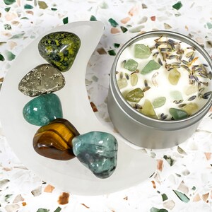 K&ouml;nnte beinhalten: Eine silberne Kerzenzinn mit einer wei&szlig;en Wachsoberfl&auml;che, verziert mit gr&uuml;nen Steinen und getrocknetem Lavendel. Eine halbmondf&ouml;rmige Schale aus mattiertem Glas enth&auml;lt verschiedene polierte Edelsteine in Gr&uuml;n-, Gold- und Silbert&ouml;nen.