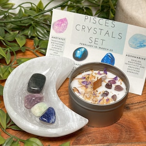 May include: A Pisces crystal set with a white selenite moon shaped dish, a silver candle tin with a wooden wick, and four crystals: amethyst, aquamarine, black tourmaline, and labradorite. The card reads "Pisces Crystals Set, February 19 - March 20".