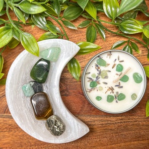 May include: A white crescent-shaped selenite dish holds various polished gemstones, including green aventurine and tiger's eye. A small candle in a silver tin is decorated with green stones and lavender. Greenery surrounds the objects on a wooden surface.
