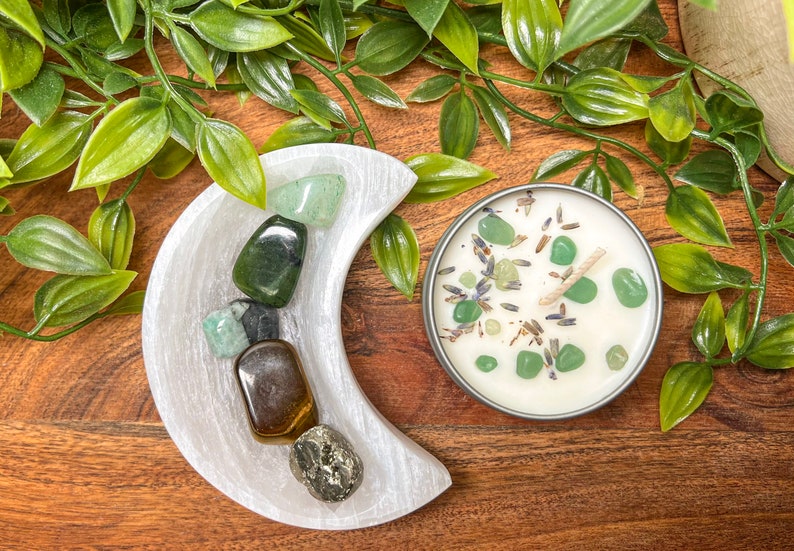 May include: A white crescent-shaped selenite dish holds various polished gemstones, including green aventurine and tiger's eye. A small candle in a silver tin is decorated with green stones and lavender. Greenery surrounds the objects on a wooden surface.