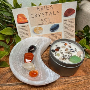 May include: A set of crystals for the zodiac sign Aries, including carnelian, citrine, hematite, and red jasper. The crystals are arranged in a white crescent-shaped dish. A candle with crystals and lavender is also included. The card reads "Aries Crystals Set March 21 - April 19".