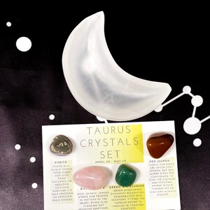 May include: A white crescent moon-shaped object sits above a card labeled "Taurus Crystals Set." The card displays four polished gemstones: pyrite, rose quartz, green aventurine, and red jasper. The background is black with white star and constellation designs.