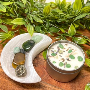 May include: A silver candle tin with a white wax candle, decorated with green stones and lavender. A white crescent moon-shaped dish holds various gemstones. Green foliage surrounds the objects, creating a natural aesthetic.