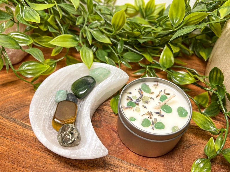 May include: A silver candle tin with a white wax candle, decorated with green stones and lavender. A white crescent moon-shaped dish holds various gemstones. Green foliage surrounds the objects, creating a natural aesthetic.