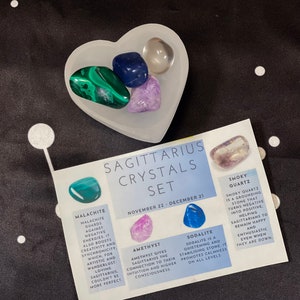 May include: A set of crystals for Sagittarius, including smoky quartz, amethyst, sodalite, and malachite. The crystals are arranged in a heart-shaped dish on a black background. The card describes the properties of each crystal.