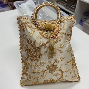 Può includere: Una pochette bianca ricamata con un motivo floreale dorato e un manico decorato con perle. La borsa ha un pendente a forma di nappa al centro.