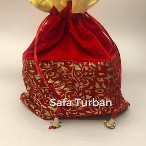 May include: Red velvet drawstring bag with gold floral pattern and gold trim. The bag has a gold ruffled top and the text "Safa Turban" printed on the front. The bag has two gold tassels at the bottom.