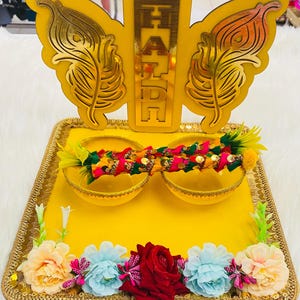 Può includere: Un vassoio giallo decorativo con bordi dorati, con un elemento a forma di farfalla e la parola "HAIR" in oro. Il vassoio contiene due ciotole gialle ed è adornato con fiori colorati e oggetti decorativi. Il design è luminoso e festoso.