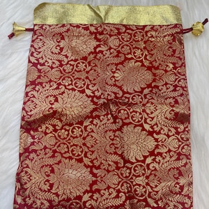 May include: A red drawstring bag with a gold brocade pattern and gold trim. The bag has a gold tassel on each side.
