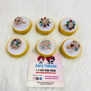 May include: Six colorful rings with rhinestones and gold settings displayed on yellow circular stands. The rings are arranged in a circle around a business card with the text "SAFA TURBAN +1-437-655-3040 www.shopsafaturban.com #safaturbanbykaran1 Follow Us On: #aainabysafaturban Unit 3-158 Kennedy Rd. South Brampton, ON L6W 3G7"