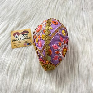 May include: A purple turban with detailed embroidery in orange, pink, and yellow, and gold sequins. The turban features an ornate design. A small card with the text "SAFA TURBAN" is next to the turban.