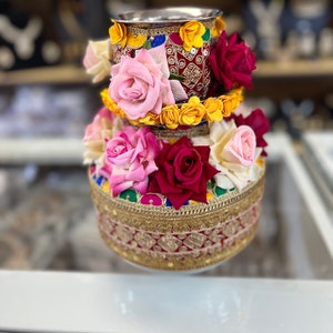 May include: A traditional Indian wedding ceremony pot decorated with colorful flowers, including pink, red, and yellow roses. The pot is adorned with intricate gold and red embroidery.