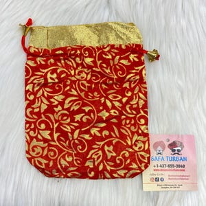 Può includere: Una borsa a cordoncino in velluto rosso con un motivo floreale dorato e un bordo dorato. La borsa è chiusa con un fiocco dorato. La borsa è appoggiata su una superficie bianca e soffice. Un biglietto da visita per Safa Turban si trova nell'angolo in basso a destra dell'immagine. La carta contiene il nome dell'azienda, il numero di telefono, il sito web e le maniglie dei social media.