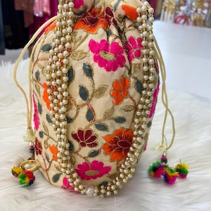 May include: Cream-colored drawstring bag with vibrant floral embroidery in pink, orange, and red. The bag is adorned with a beaded pearl strand and colorful tassel accents. The bag's design features gold thread accents.