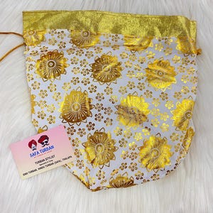 May include: A white drawstring bag with a gold floral pattern and a gold trim. The bag is made of a sheer fabric and has a gold drawstring closure. The bag is sitting on a white surface. The bag has a business card attached to it that says "Safa Turban, Turban Stylist, Specialist in Tying, Sikh Turban, Hindu Turban (Safa), Thalapa."