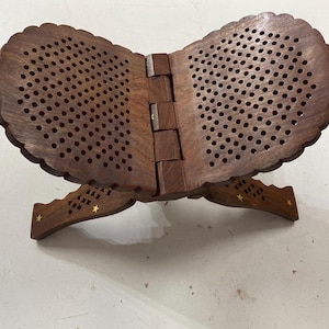 May include: A dark brown wooden book stand. The stand is foldable and features a decorative, scalloped edge and a pattern of small holes. The legs have a star-shaped design. It is designed to hold a book open.