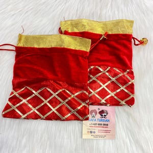Może przedstawiać: Dwa czerwone aksamitne woreczki ze sznurkiem, ze złotymi wykończeniami i złotym wzorem w romby. Woreczki mają złoty pasek na górze i małą złotą koralik na sznurku. Widoczna jest wizytówka z napisem "SAFA TURBAN".