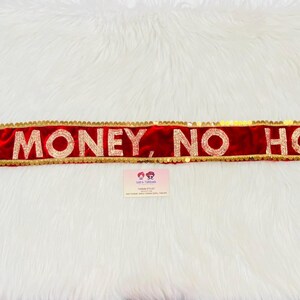 Puede incluir: Una faja roja con ribete dorado y las palabras "NO MONEY, NO HONEY" en letras doradas. La faja tiene lazos trenzados coloridos en cada extremo. La faja se muestra sobre una superficie blanca y esponjosa.