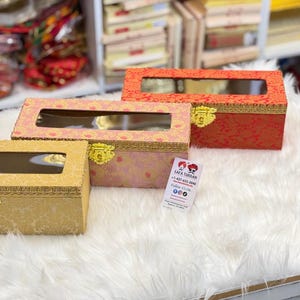 May include: Three decorative rectangular boxes with clear viewing windows. The boxes are in shades of gold, pink, and red, with gold clasps and trim. They sit on a white, fluffy surface, suggesting a display of gift boxes or storage containers.