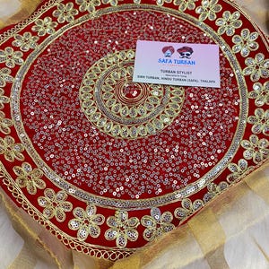 Puede incluir: Una bandeja de turbante de terciopelo rojo con bordados dorados y lentejuelas plateadas. La bandeja está decorada con un patrón circular de flores y tiene una tarjeta de visita que dice "Safa Turban, Estilista de turbantes, Especialista en atar, Turban sikh, Turban hindú (Safa), Thalapa".
