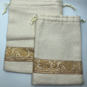 May include: Two beige burlap drawstring bags with gold trim. The bags have a woven texture and feature a decorative gold floral pattern.