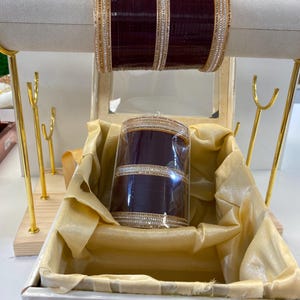 Può includere: Un set di tre braccialetti viola con decorazioni dorate e bianche. I braccialetti sono in una scatola bianca con un bordo dorato e una fodera in raso giallo. La scatola ha un coperchio trasparente e il numero 2,4 è visibile nell'angolo in basso a destra.