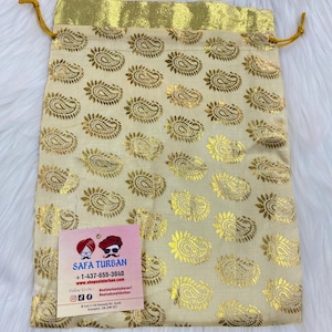 May include: A gold drawstring bag with a paisley pattern. The bag is made of a soft, silky fabric and has a gold trim. The bag is perfect for storing small items, such as jewelry or gifts. The bag has a tag attached to it with the text "Safa Turban" and contact information.