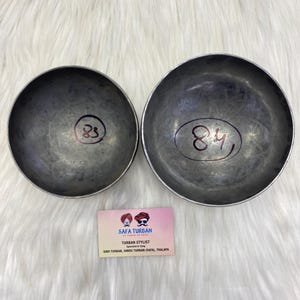 Pode incluir: Duas tigelas de metal prateado com os números 83 e 84 escritos nelas. As tigelas estão sobre uma superfície branca e felpuda. O texto "SAFA TURBAN TURBAN STYLIST Especializado em amarrar SIKH TURBAN, HINDU TURBAN (SAFA), THALAPA" está em um cartão branco abaixo das tigelas.