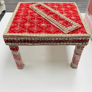 May include: A small, square red table with ornate gold and silver trim. The table has four legs wrapped in a matching red fabric. A rectangular piece of fabric with similar embellishments rests on top.