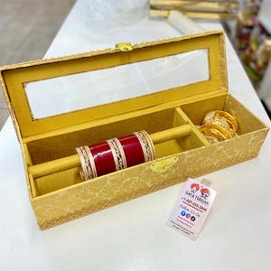Puede incluir: Una caja dorada con tapa transparente, diseñada para guardar joyas. La caja tiene un interior de terciopelo y un compartimento para pulseras. La caja está decorada con intrincados bordes dorados y un patrón floral. La caja está etiquetada con "Safa Turban +1-437-635-3040 Síguenos en:"