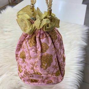 Puede incluir: Un bolso rosa con cordón y bordado floral dorado. El bolso presenta detalles de tela dorada y asas de cuentas. El bolso se utiliza probablemente para llevar objetos pequeños y tiene un diseño decorativo.