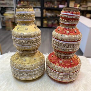 Peut inclure: Deux vases décoratifs ornés. L'un est doré, l'autre rouge avec des motifs floraux dorés. Les deux présentent des perles et des embellissements complexes. Ils sont présentés sur une surface blanche et moelleuse, mettant en valeur leurs motifs élaborés.