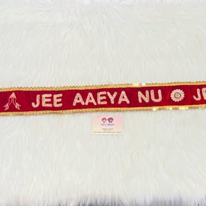 Può includere: Un nastro di velluto rosso con bordi dorati e paillettes. Il nastro ha il testo "AAO JI JEE AAEYA NU JEEJA J" ricamato in bianco.